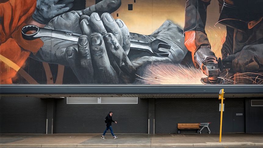 A huge wall mural of hands holding spanners and welding. Beneath it a slightly blurred man is walking.