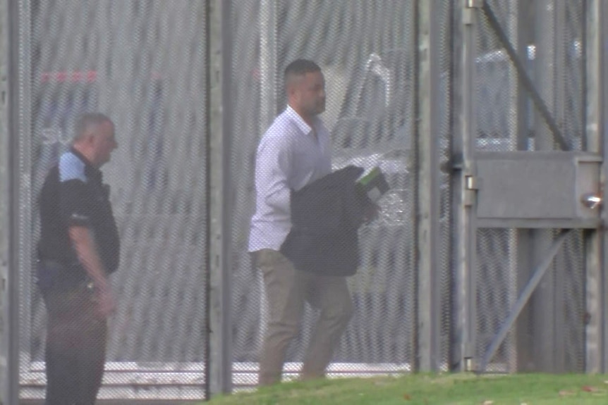 A man in a white short holding a piece of clothing leaves a prison with a man following him from behind.