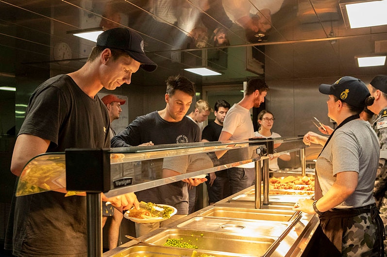 Three men are seen in the process of loading food onto their plates, with a queue of people behind them. Navy workers on right.