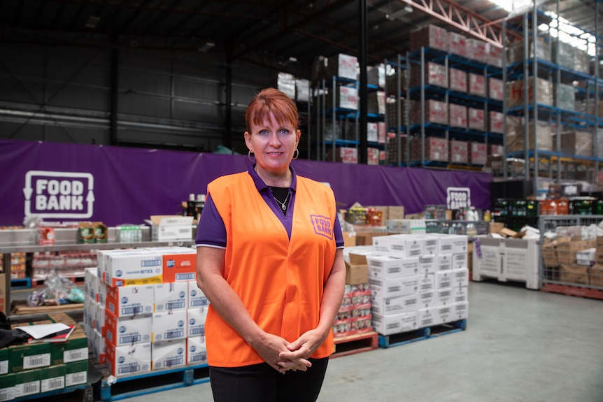 A woman wearing an orange high-vis vest stands in front of stacks of food.