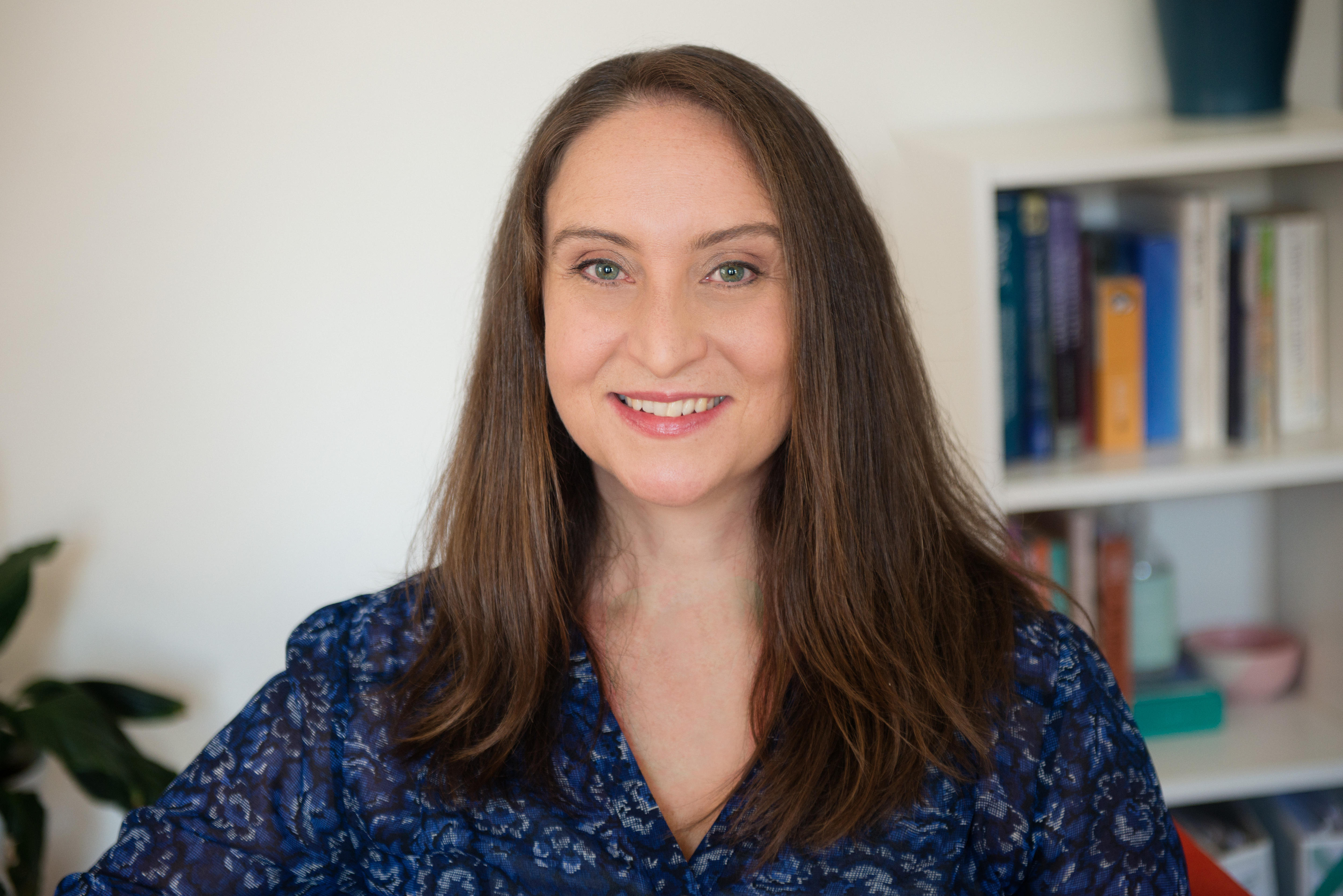 A woman smiles. She has long, straight brown hair, a blue outfit, and a bookcase behind her.