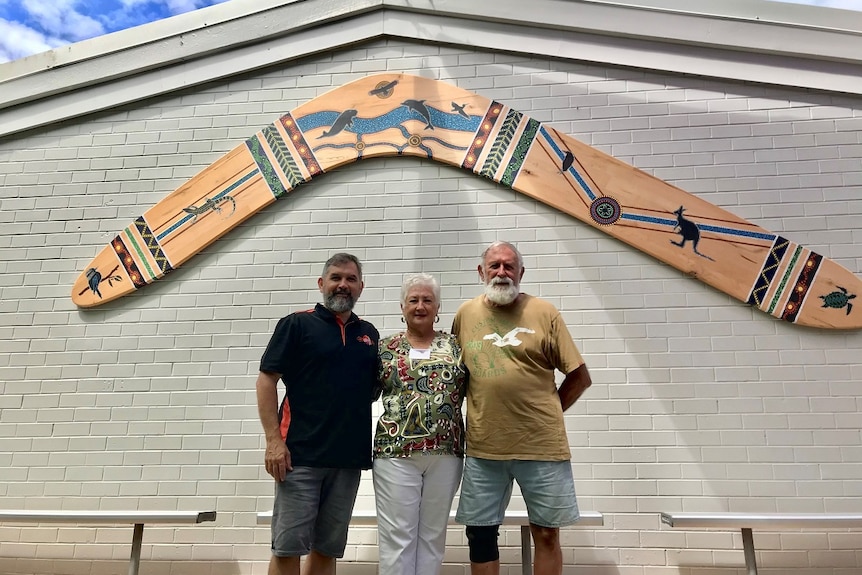 Three people standing underneath a large boomerang wall feature on the side of a building under a blue cloudy sky