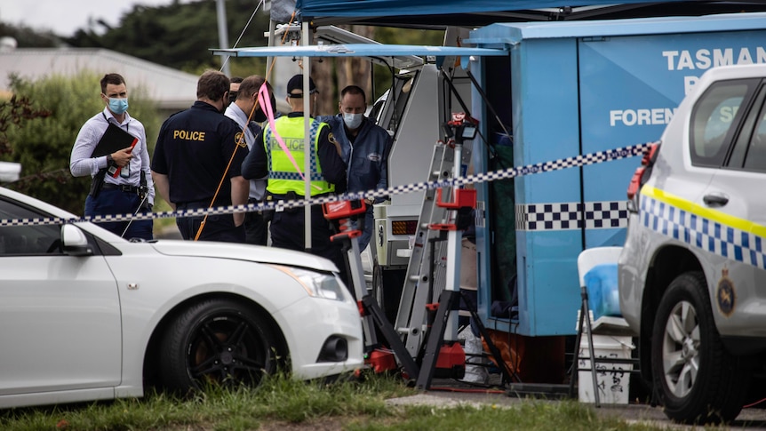 Police officers stand around near a forensics tent with a police car in front. 