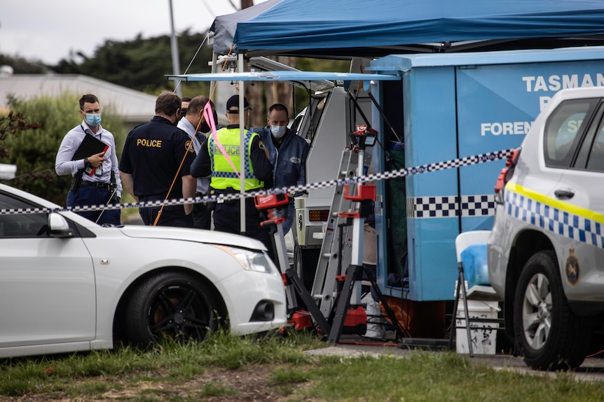 Police officers stand around near a forensics tent with a police car in front. 