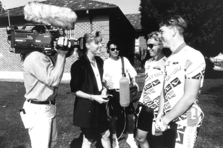 Black and white photo of Tighe and cameraman interviewing cyclists.