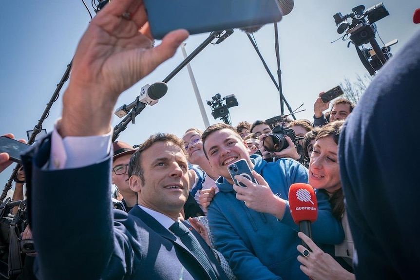 Emmanuel Macron holds up a phone as he poses with a crowd of people