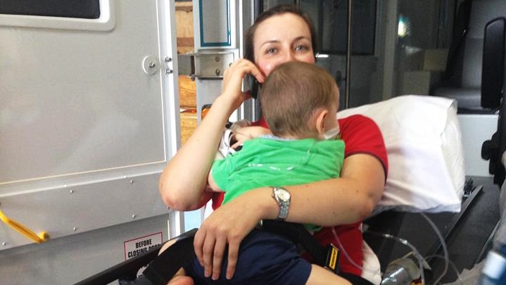 A midwife on an ambulance stretcher cradles a toddler while on the phone