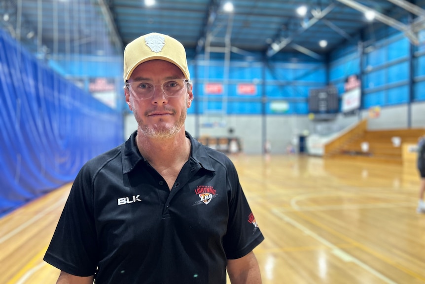 A man with a hat and black top looking at the camera while standing on a basketball court.