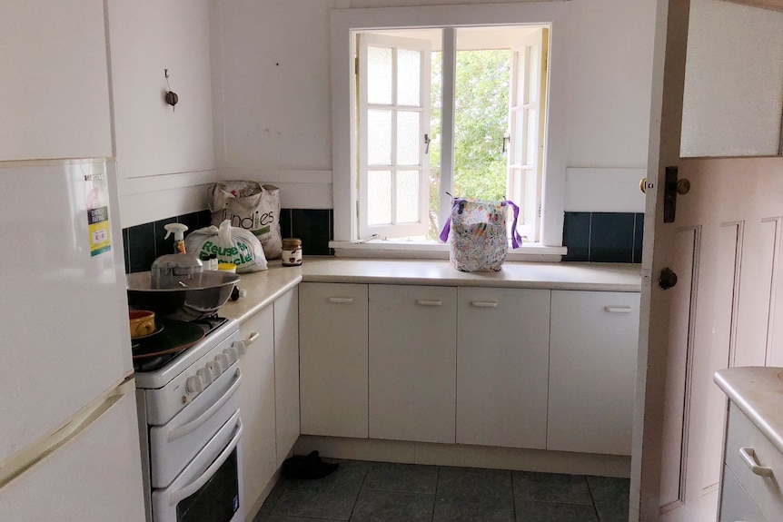 A run-down kitchen with cream cupboards and benchtops is seen with a blue tiled floor below. The kitchen windows are open.