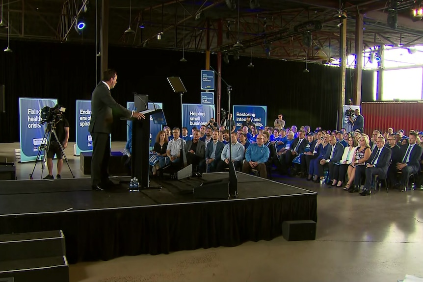 People in blue watch Matthew Guy deliver a speech, with Liberal signage in the background.