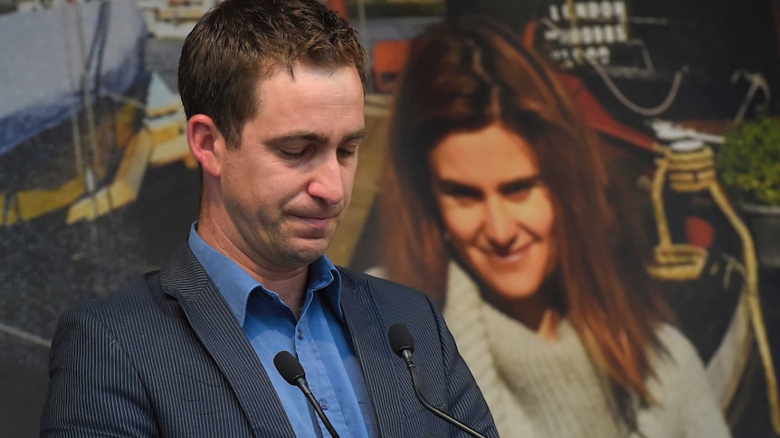 Brendan Cox stands at a microphone looking down, with an image of Jo Cox behind him