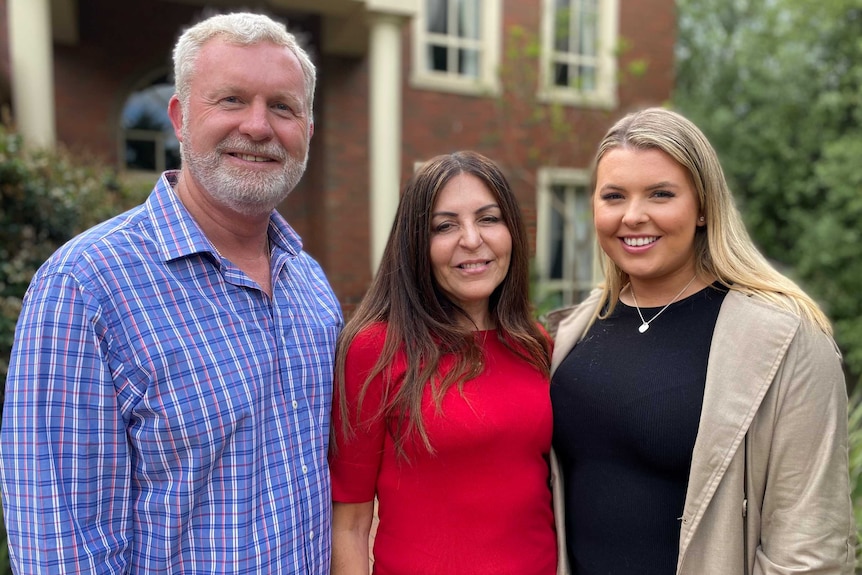 A man with grey hair and a beard stands with a dark haired woman and her blonde daughter.