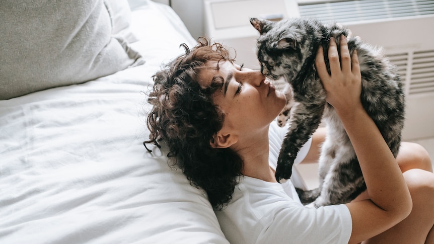 A woman with brown curly hair sits on the floor next to a bed made with white linen, kissing her cat on the nose