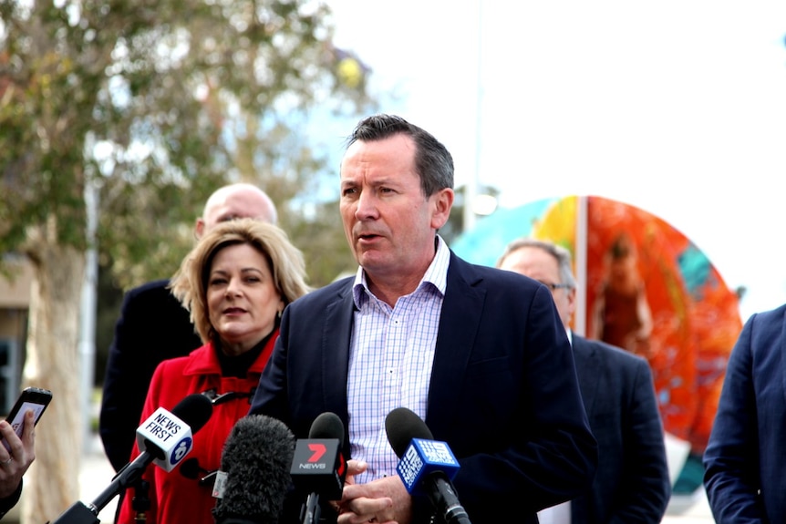 WA Premier Mark McGowan stands at a microphone flanked by a woman wearing a red jacket, with some people standing nearby.