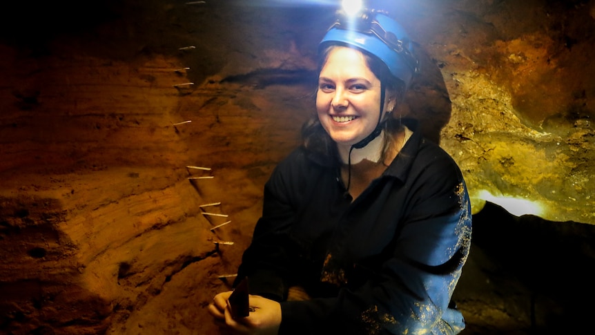 A woman wearing a helmet with headtorch holds a trowel inside a small cave