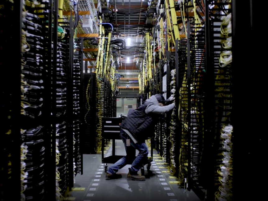 A man leans to work on a large server, standing in an aisle of servers with cables running everywhere.