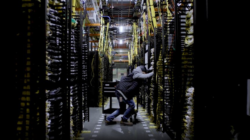 A man leans to work on a large server, standing in an aisle of servers with cables running everywhere.