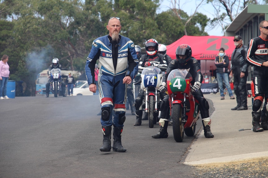 A man in blue racing leathers walking with racers on motorbikes behind him