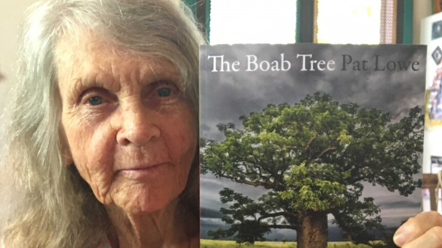 A woman holds a book showing the cover of a boab tree in leaf beneath a stormy sky