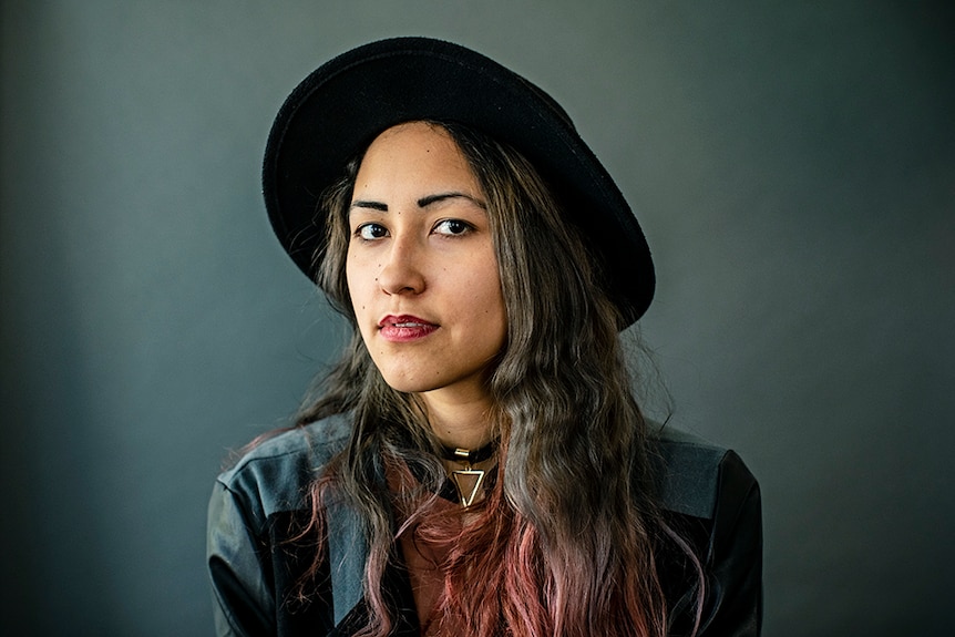 Anchuli Felicia King wears black hat and jacket and poses in front of grey background.