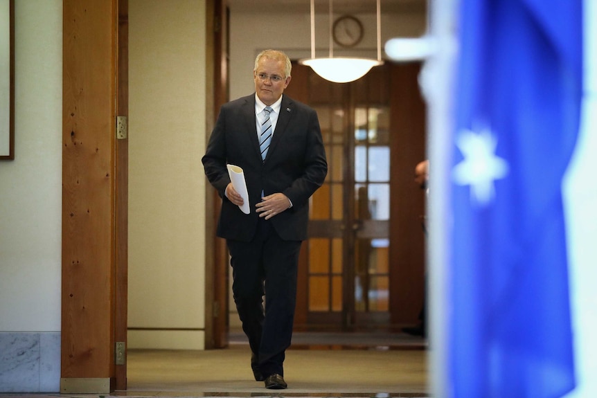 A man in a dark suit walks towards the camera carrying folded papers.