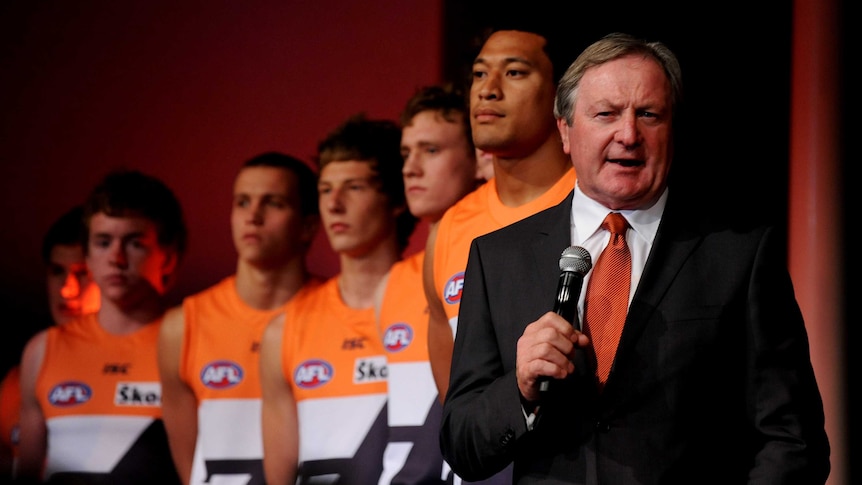 An AFL coach stands on the stage with his players behind him.