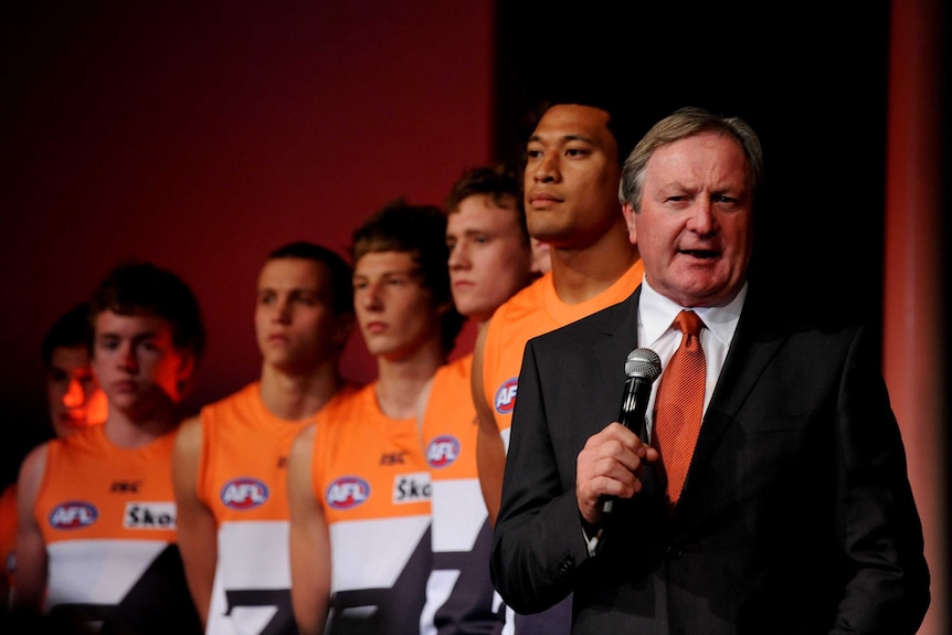 An AFL coach stands on the stage with his players behind him.
