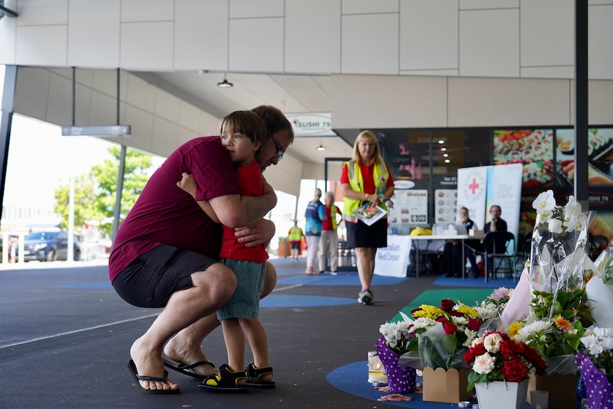 a man hugs a young boy in front of a pile of floral tributes