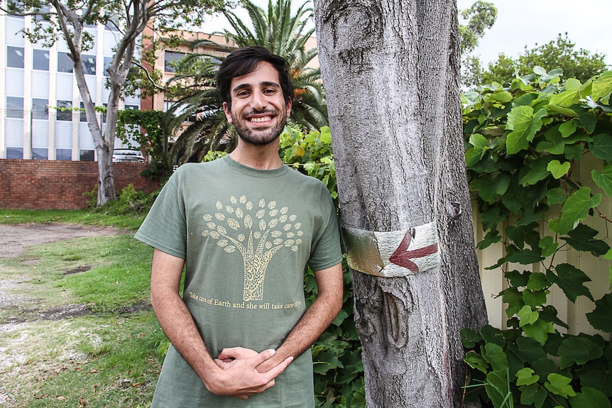A young man wearing a green t.shirt depicting Arabic calligraphy shaped like a tree, stands underneath a tree.