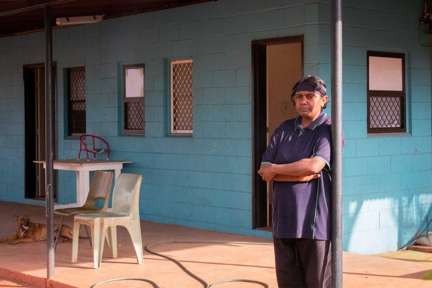 An Indigenous woman stands in front of a basic dwelling.