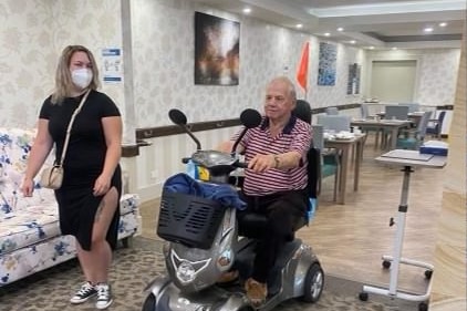 An elderly man rides a mobility scooter through a room of a nursing home with a younger woman walking next to him.