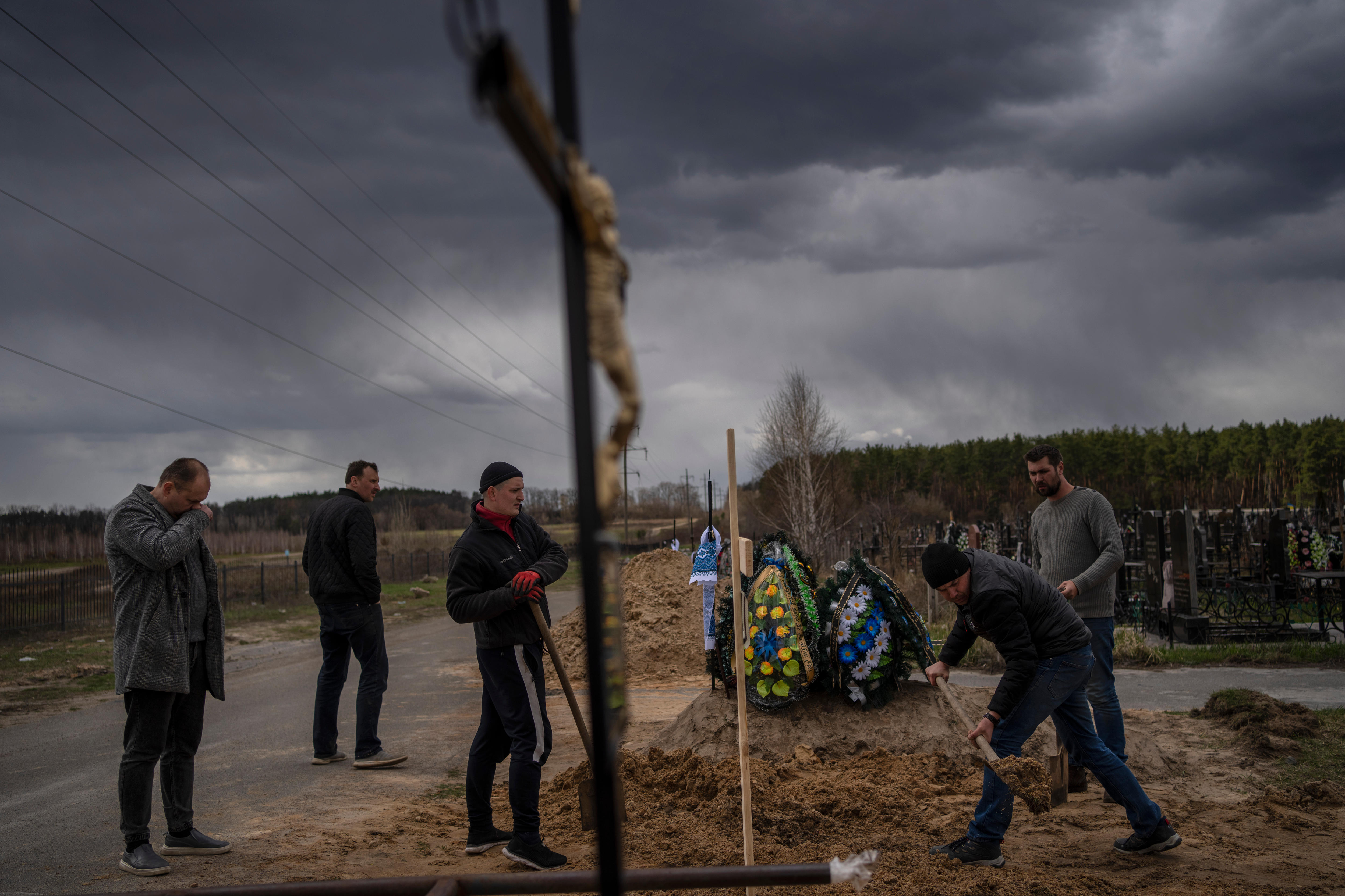 Men stand with shovels near wreaths of flowers with dark clouds overhead.