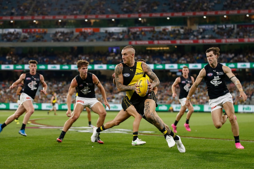 Dustin Martin carries the ball while running. He's surrounded by four Carlton players