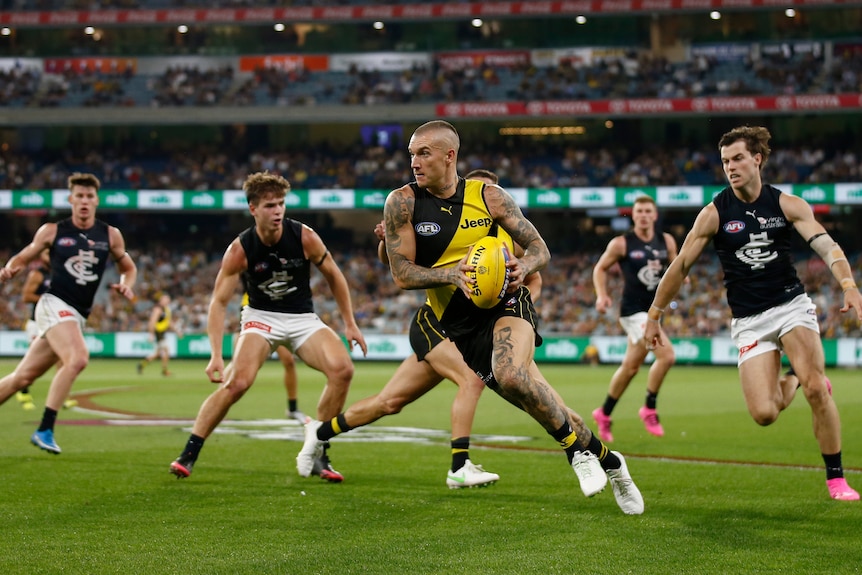 Dustin Martin carries the ball while running. He's surrounded by four Carlton players