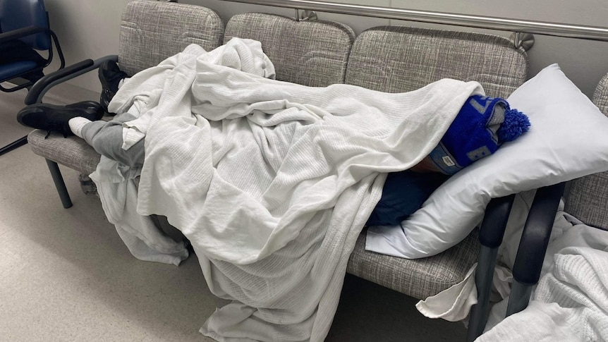 Boy laying down across 3 seater couch in hospital hallway. He is covered by a messy blanket and is wearing black runners.