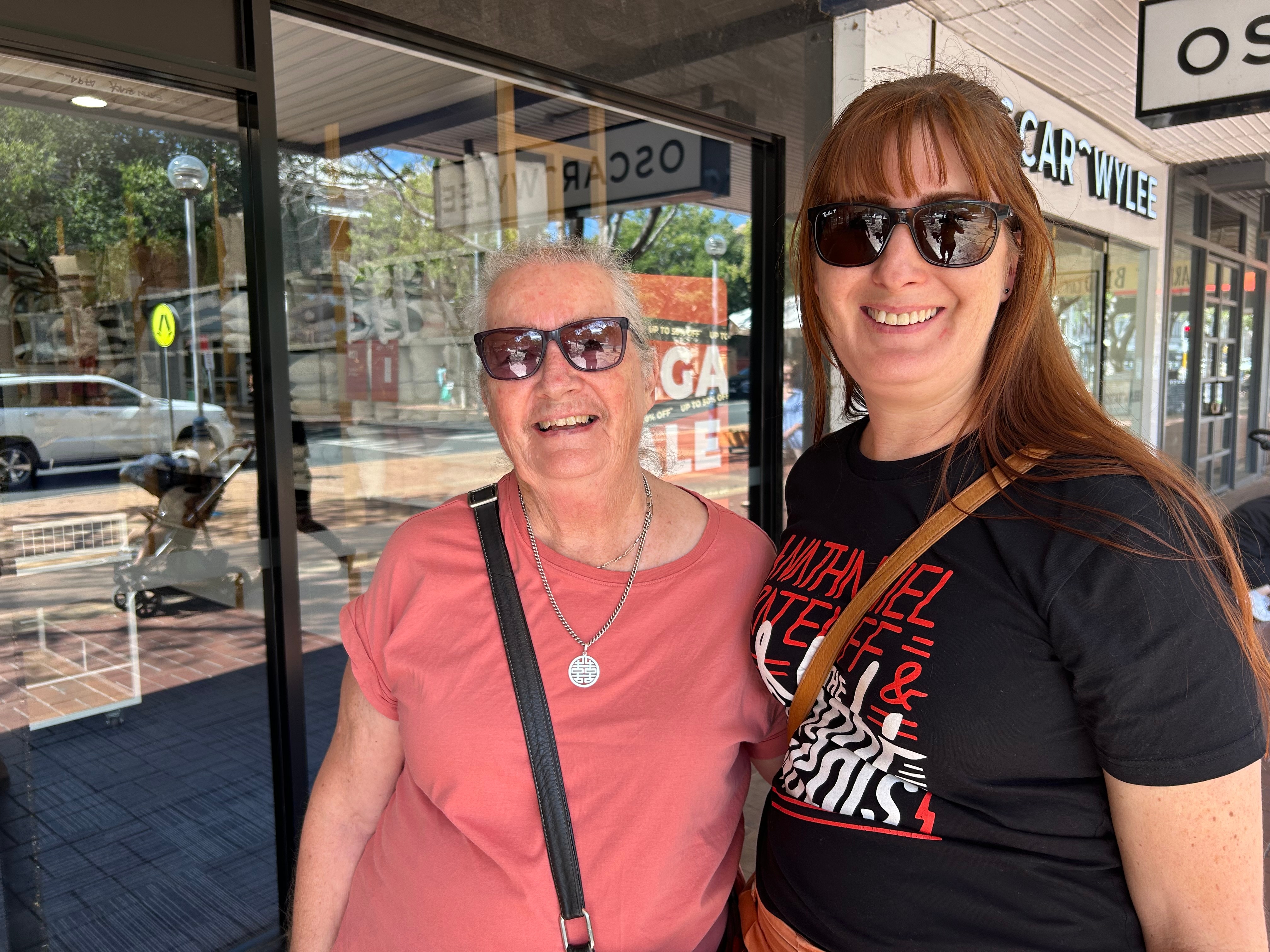 Una mujer joven con cabello castaño y una camiseta negra está junto a su madre con una camiseta rosa.