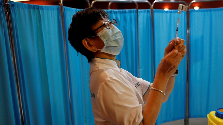 A nurse in a surgical mask holds up a syringe with a hospital curtain in the background.