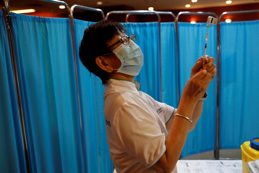 A nurse in a surgical mask holds up a syringe with a hospital curtain in the background.
