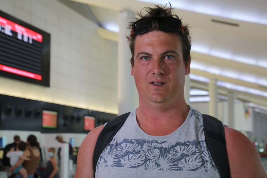 A man carrying a backpack stands in Perth Airport with a departures board in the background.