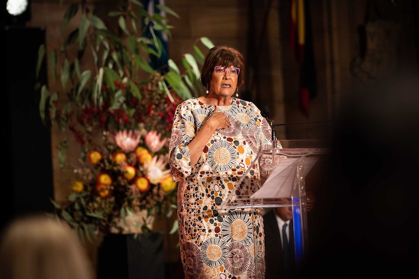 Pat Turner is standing at a podium speaking. She is wearing pink-framed glasses and a dress with an Aboriginal art design.