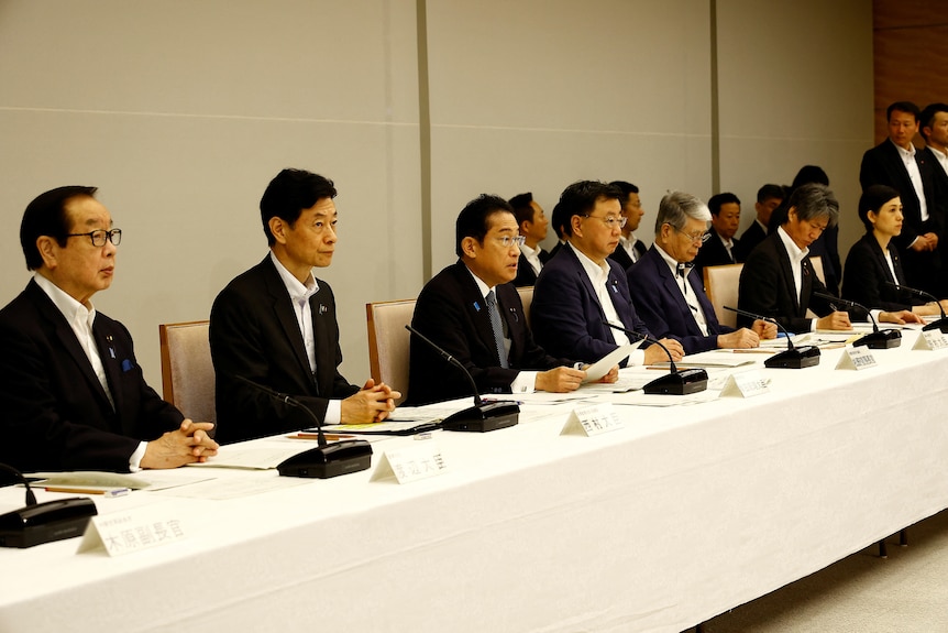 A long table with several menn in black suits sitting on one side with microphones and placards in front
