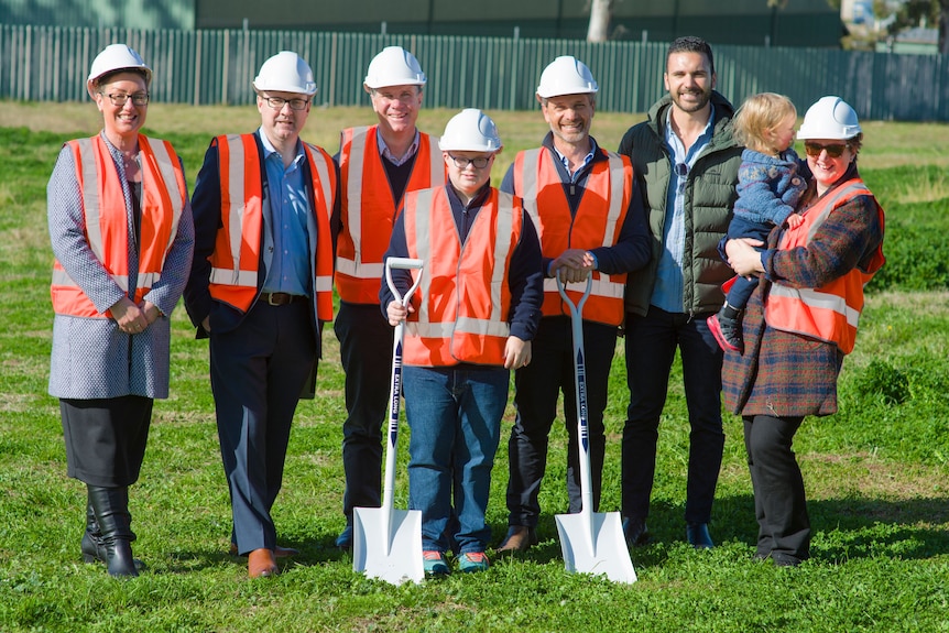 Eight individuals standing in an empty lot of land with several shovels in the ground
