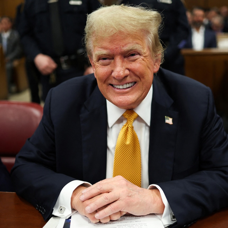 Trump smiles in a yellow tie and suit in a court