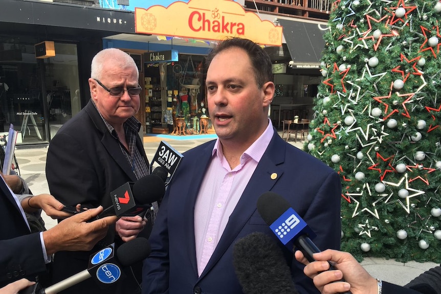 Philip Dalidakis stands in front of a Christmas tree and speaks into media microphones.