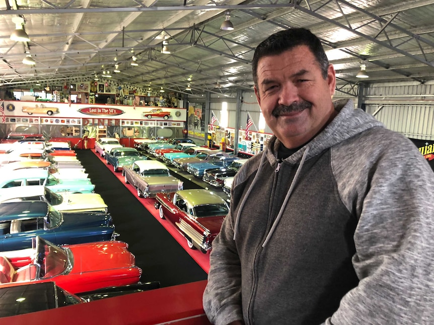 A man in a grey hooded sweatshirt stands above around 100 classic cars of different colours.
