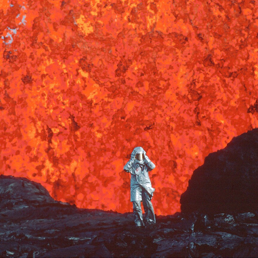 A volcanologist in aluminized silver suit stand on hardened molten rock in front of a lava burst with hands held up to helmet.