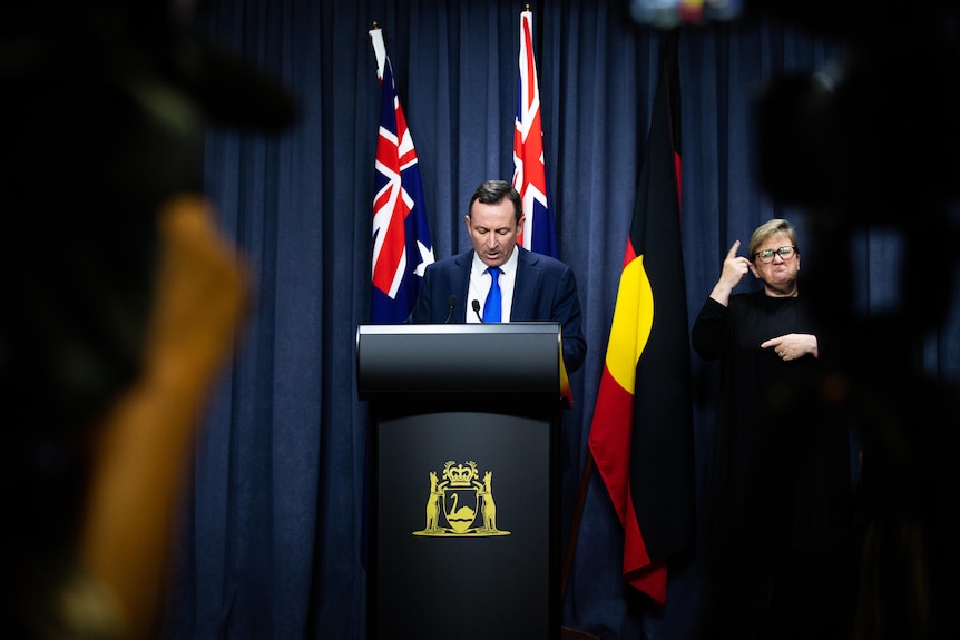 Man looks at notes on lectern as he speaks in front of the Australian, Western Australian, and Aboriginal flags.