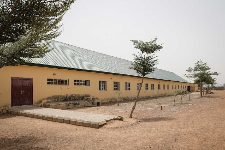 A long single storey building is seen with small trees planted in a row running along its side.
