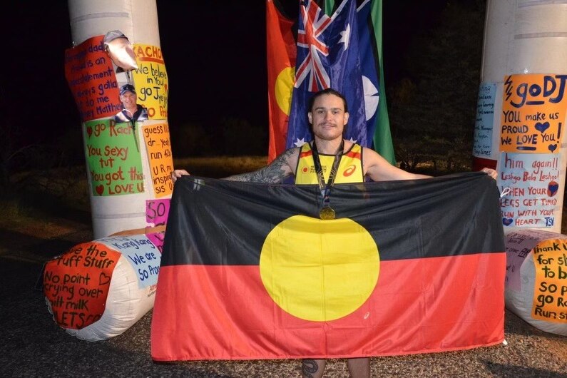 Derrick Cusack stands smiling at a running finish line holding the Aboriginal flag open in front of him. 