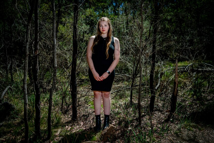 A woman with long brown hair, in black dress stands in partially burnt out bush, stark black and green colours. 